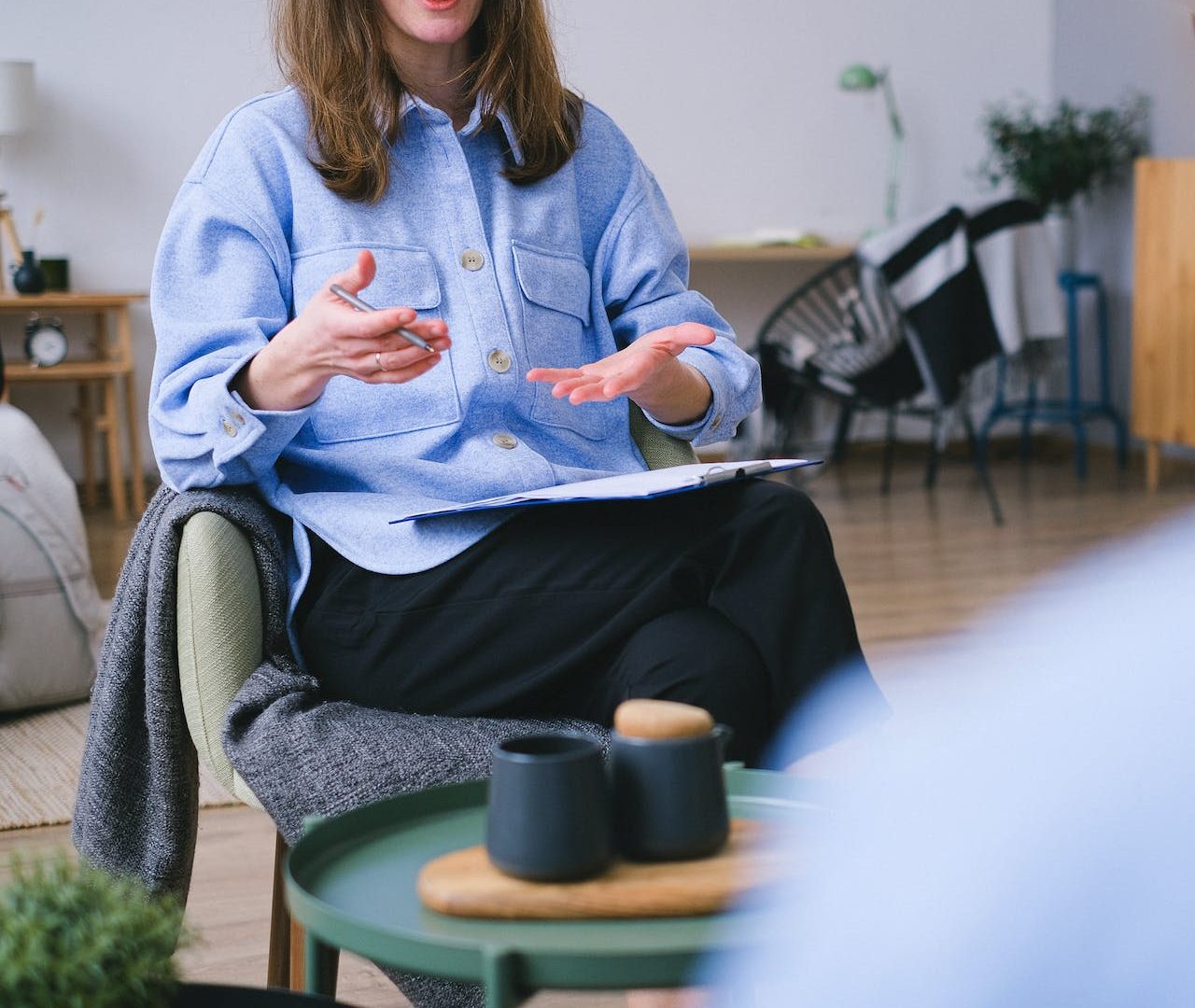 a woman with medium length brown hair in a blue shirt is speaking and gesturing with her hands with a notepad balanced on her knee