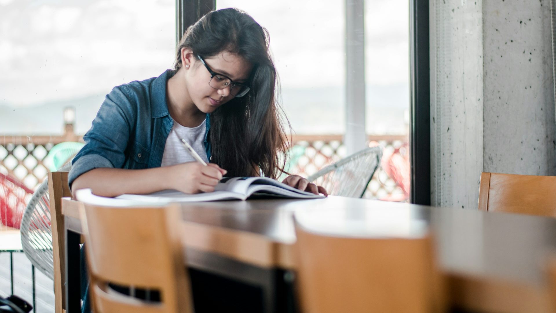 A young woman with long black hair and thich black rimmed glasses is sitting at a long table alone writing in a large exercise book.