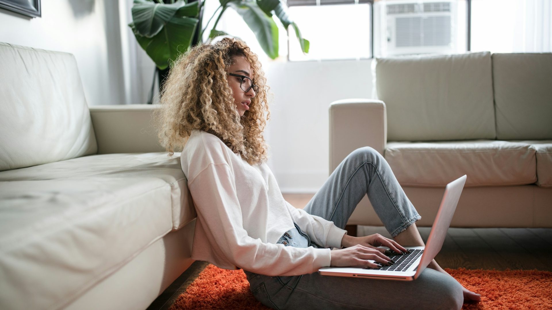 a young woman with curly hair and glasses sits on the floor on an orange carpet reading something on her laptop while leaning against a sofa