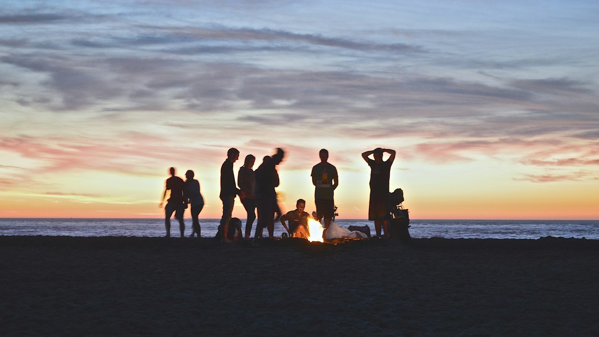 A group of people sat and stood around a fire on a beach as the sun is rising.