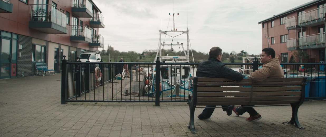 Two people sitting on a bench chatting at a marina with flats around them a boat is visible in the water behind them.