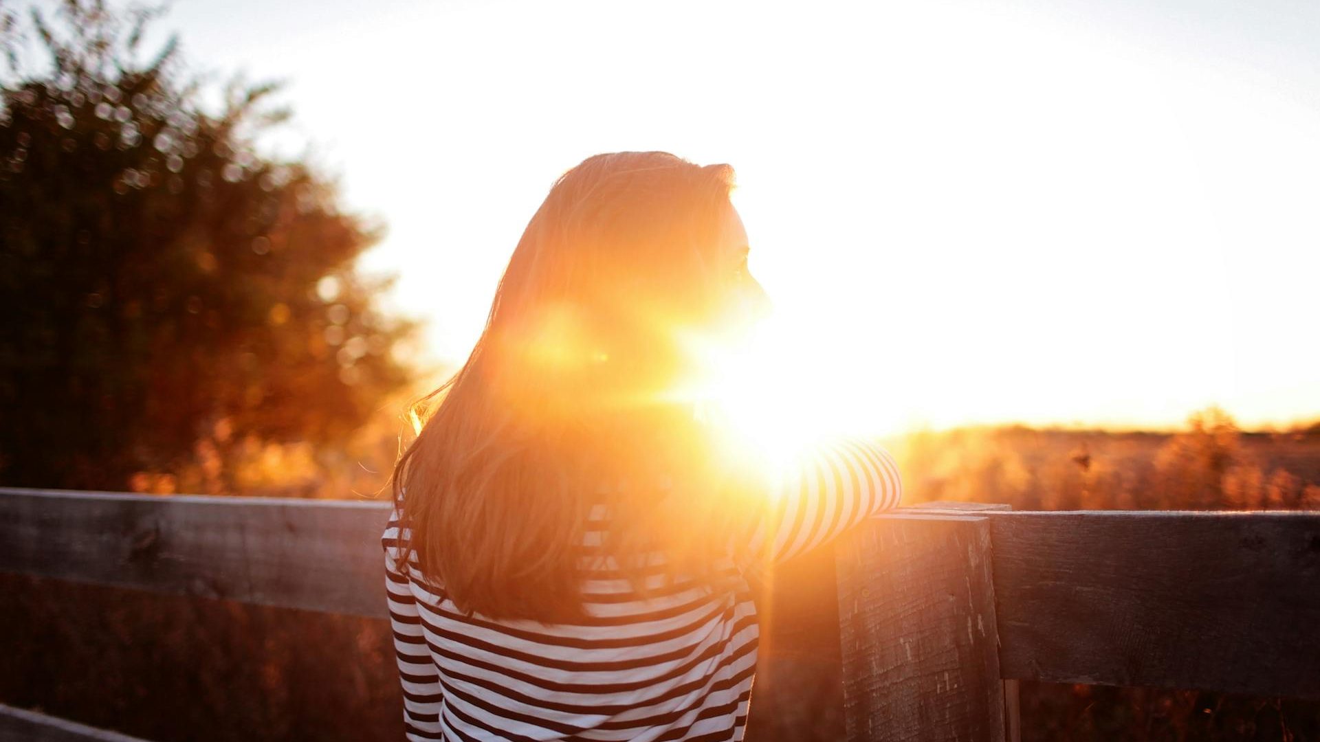 the silhouette of a woman with the sun behind her head. She has long brown hair and a striped tshirt on and is leaning against a wooden fence.