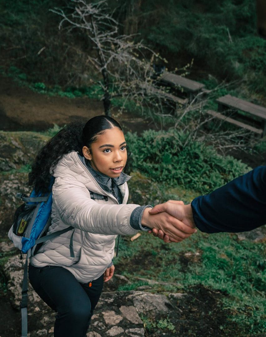 a young black woman with long black hair tied back, wearing a light down jacket, hiking in nature accepting a hand up to climb something