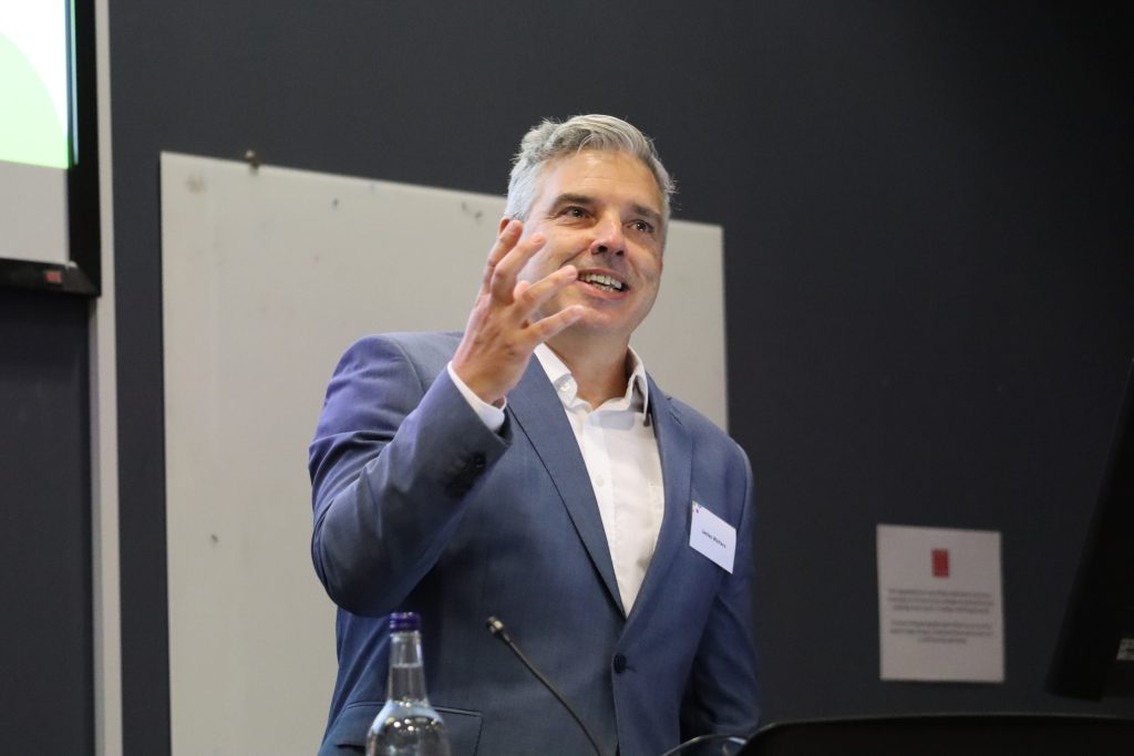 Professor James Walters speaking at the lecturn for the Hub event. He is white with short grey hair and is wearing a steel blue suit with a white shirt.