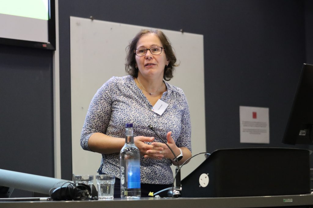 Tania Gergel speaks at the Hub launch, she wears a short sleeved top and has short dark hair and glasses.