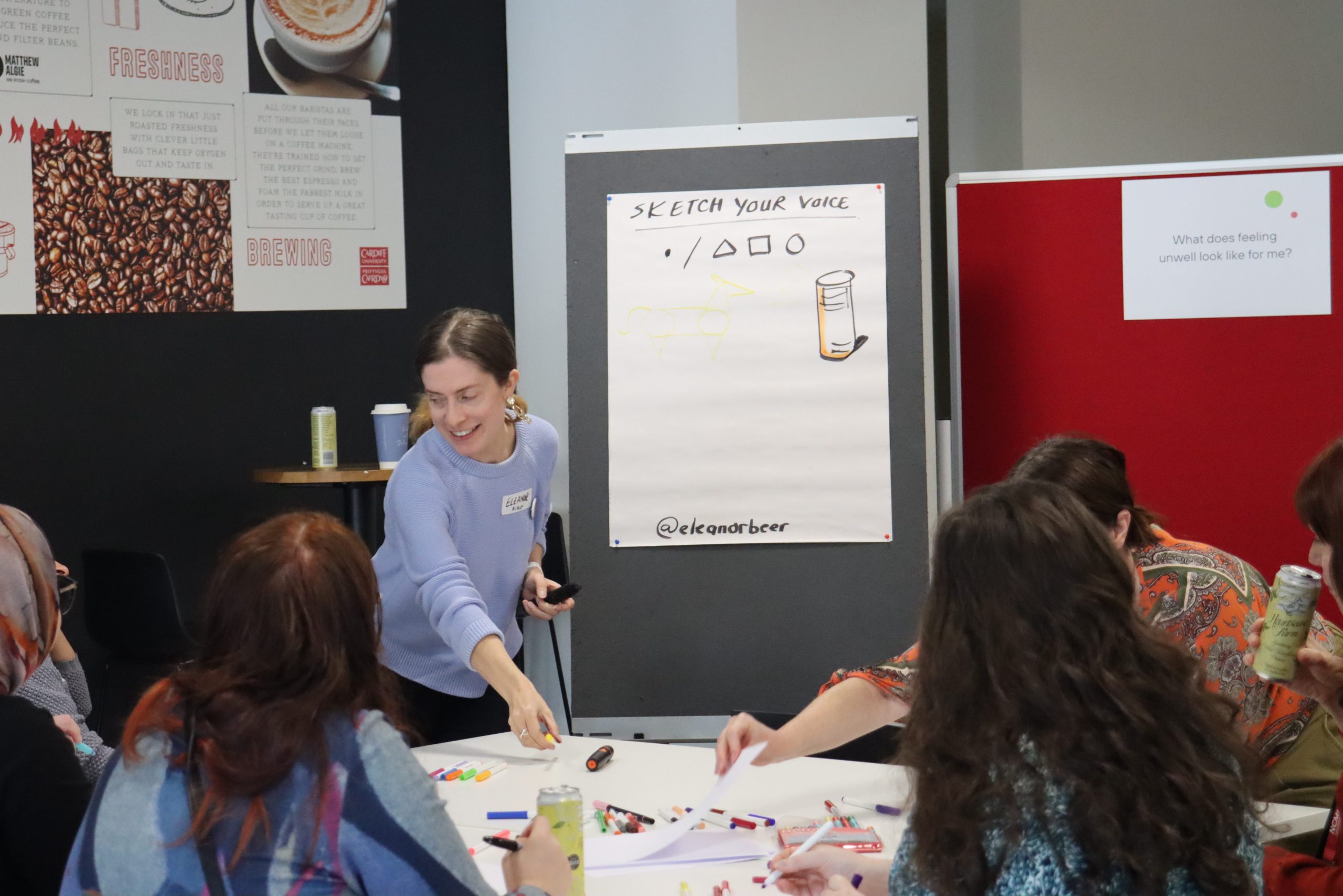 A young woman speaks to a table of people which has lots of drawing materials. 