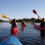 Three people kayaking on a lake in red kayaks. THere are two woman closer to the camera, facing away with brown and blonde hair and they have their oars raised. There is a man in front of them also facing away and he is wearing a light shirt and dark cap.
