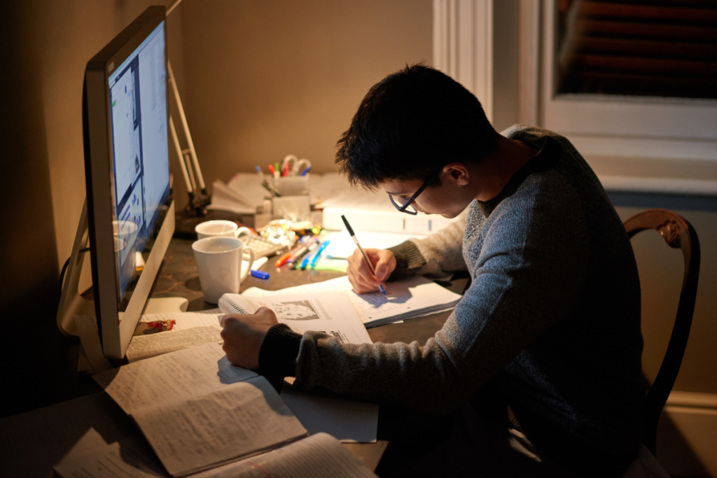 A young man with olive skin and dark hair and black glasses sits leaning over a desk as he makes notes in a book with a screen of text in front of him. 