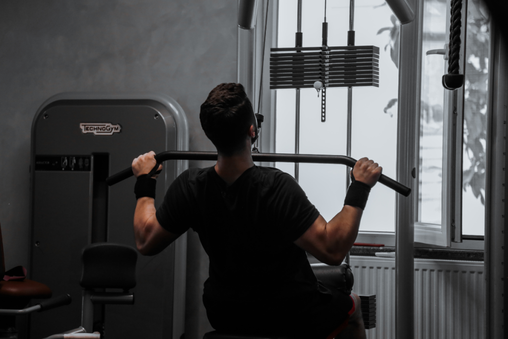 A young white man with dark hair uses a weight machine at a gym. He wears a dark shirt and wrist cuffs and is pulling a bar down towards his chest.