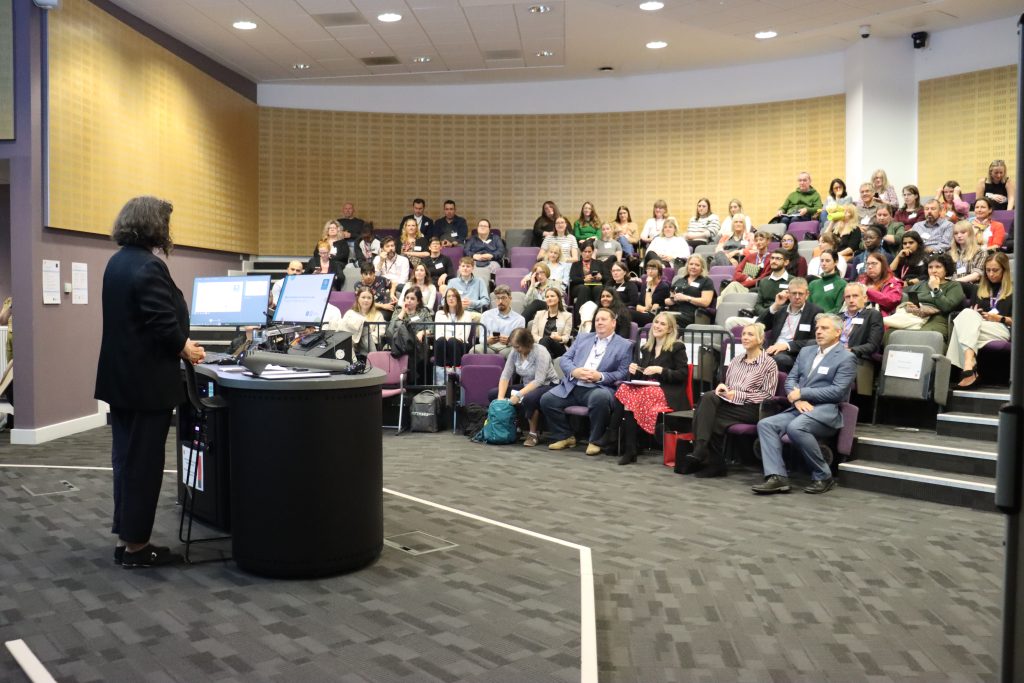 Professor Wendy Larner wearing a dark suit and shoulder length dark hair speaks to a busy lecture theatre as she opens the Hub event.