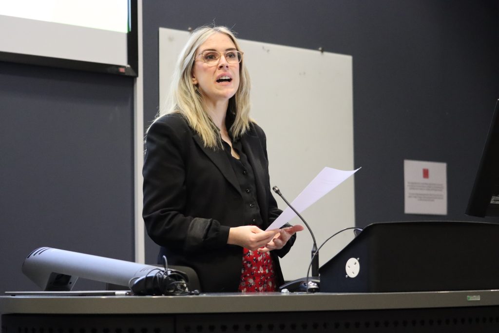 Sarah Murphy MS speaks to the audience of the Hub event while standing at the lecture theatre podium. She has long blonde hair and glasses, and wears a dark jacket with a red skirt.