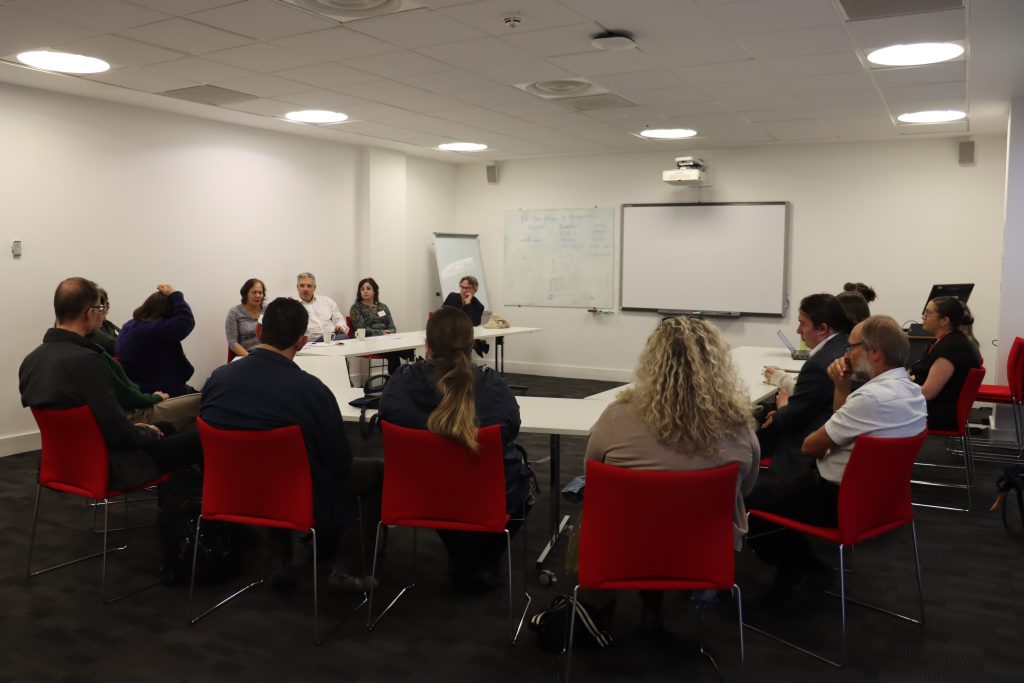Around 15 people sitting on red chairs in a 'u' shape all taking part in a workshop.