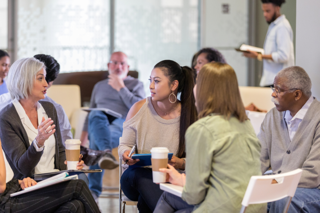 A room busy with groups of people from a mix of ethnicities and ages. Closest to the camera an older white woman with a grey bob haircut speak to a young asian woman with long black hair and older black man with short cropped grey hair, moustache and glasses.
