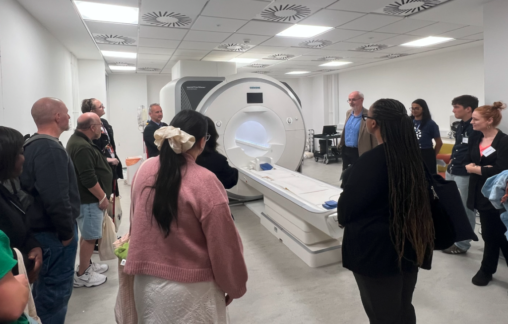 Visitors of the Hub event on a tour with Professor Neil Harrison who is speaking next to an MRI machine. A large circular white machine with a bed sticking out the front. Around ten people are watching Neil speak. 